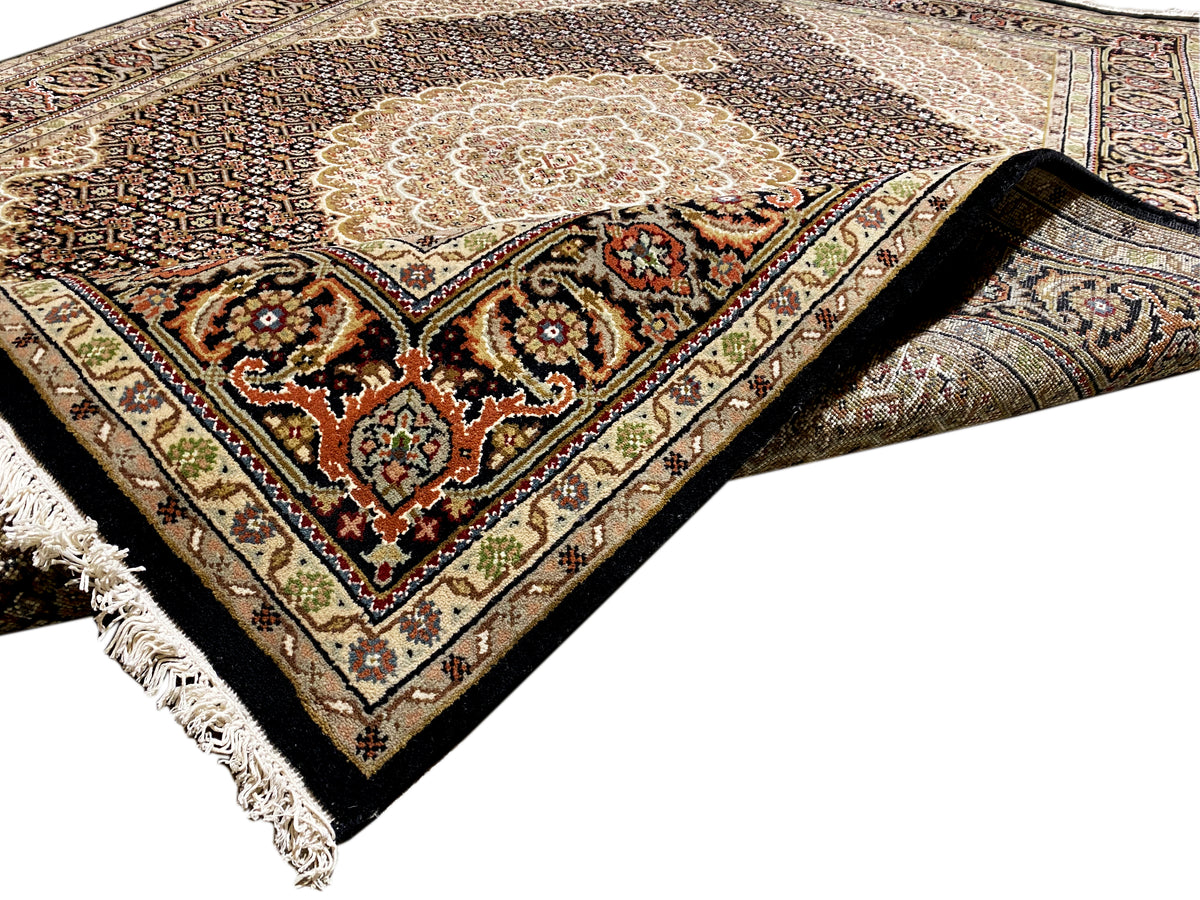 Teppich in Ecke: beiges Medaillon, dunkles Feld mit Ornamenten, Zierbordüre und Fransen.