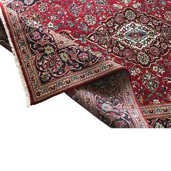 Red patterned rug with a border and fringe along the edge, fabric visible on one side