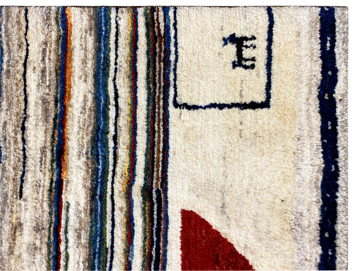 Gabbeh-Teppich Detail: cremefarbene Fläche mit blauen Linien, rotem Dreieck und Signatur-Motiv.