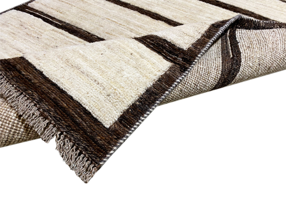 Corner view of the kilim rug: striped pattern in beige, dark lines, edge with fringes.