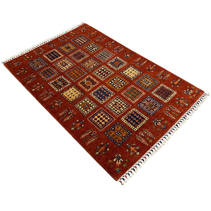 Brick-red wool rug with a geometric tile pattern and fringes at the corners, angled.