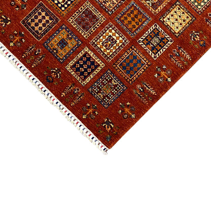 Corner area: brick-red wool rug with tile ornaments and white fringes, rectangular.