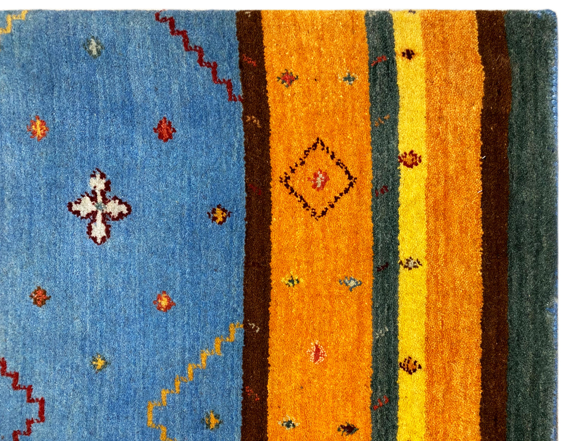 Loom Luri Teppich Detail: blaues Feld mit kleinen floralen Motiven, daneben orangefarbene Streifen.