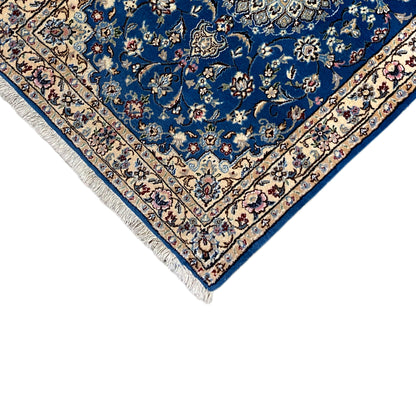 Corner of a blue Nain rug with a floral border and fringes at the tip.