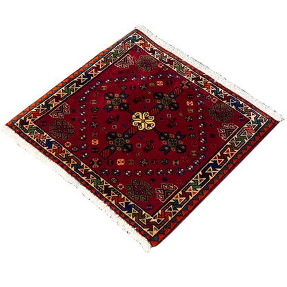 Red square Poshti rug with a symmetrical pattern, multicolored decorative border, and fringes