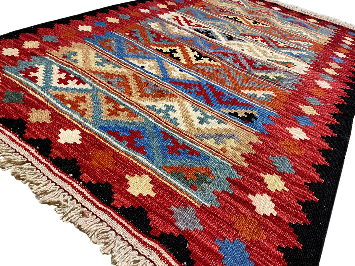 Close-up of a kilim rug: geometric patterns in red, blue, and beige, with fringes along the edge.
