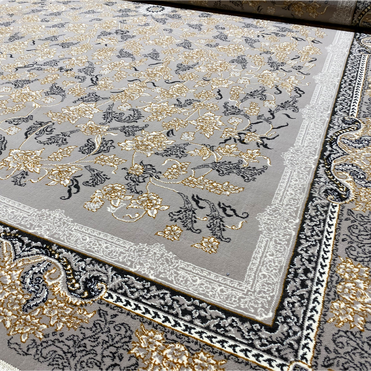 Close-up of a carpet edge with decorative trim, gold-colored floral ornaments, a gray background, and fringe