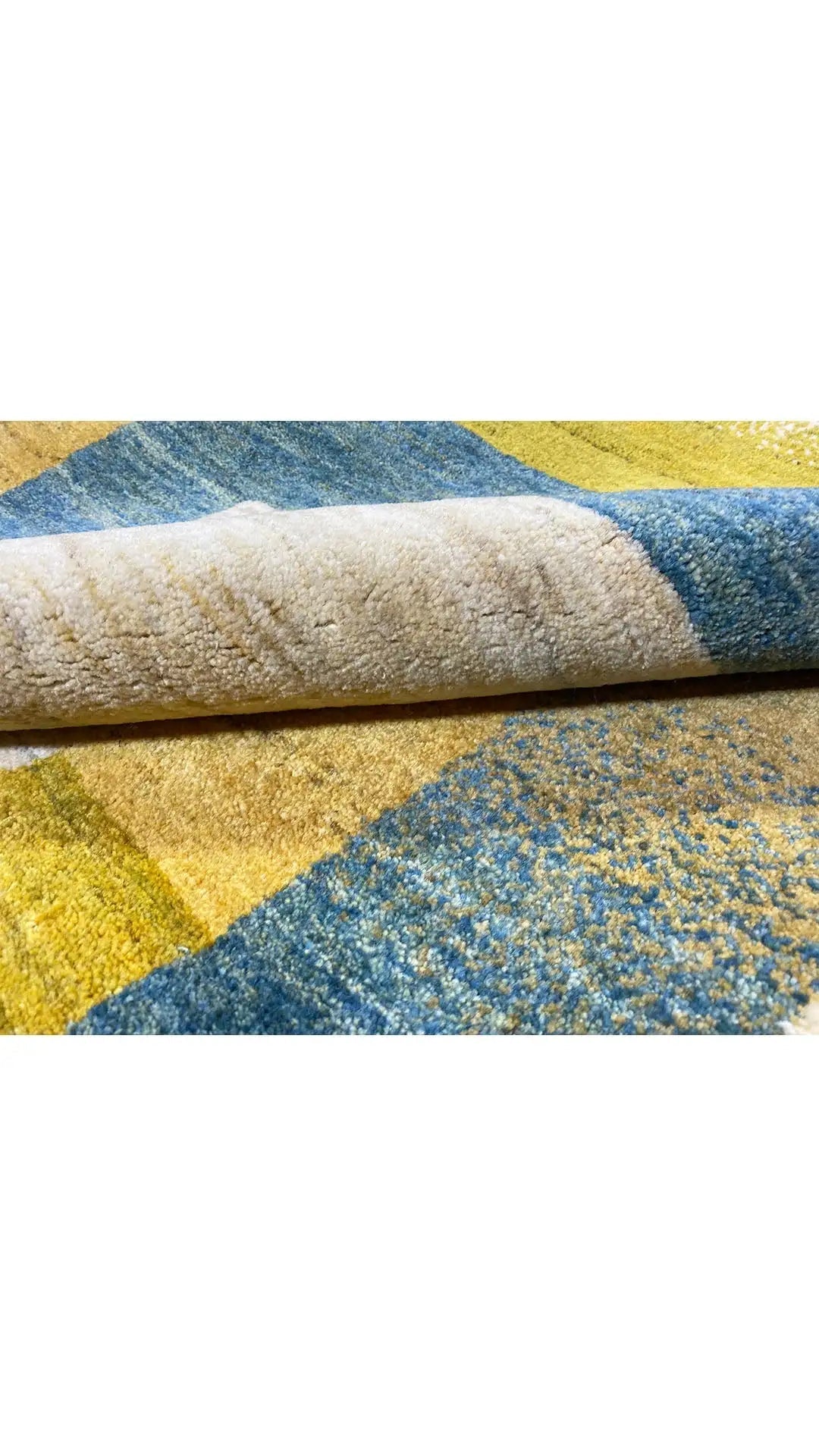 Gabbeh rug rolled at the edge: blocks of blue, yellow, and beige with a fine texture.