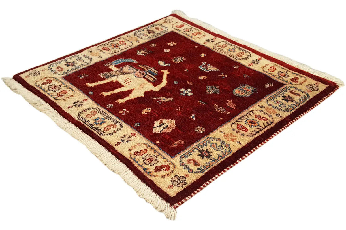 Square burgundy rug with an elaborately decorated border and fringes
