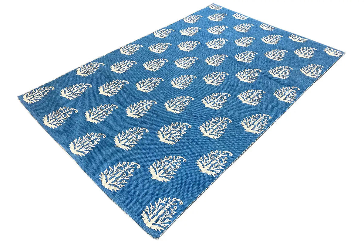 Cream-colored kilim with blue tree/branch motifs, rectangular, photographed diagonally.