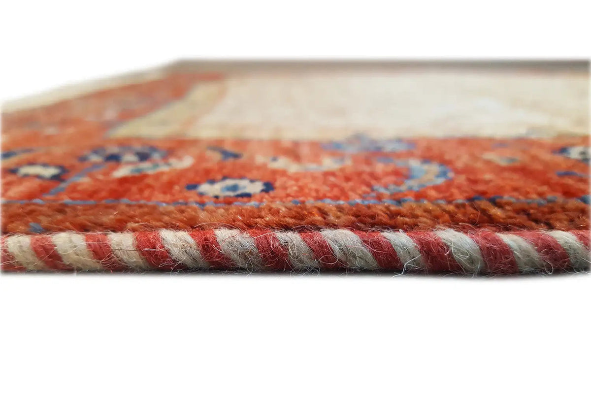 Close-up of a carpet edge: red-and-beige ornamental pattern, with rolled-up pile and fringes.