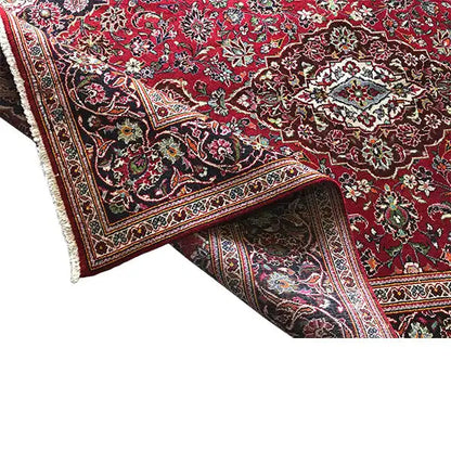 Red patterned rug with a border and fringe along the edge, fabric visible on one side