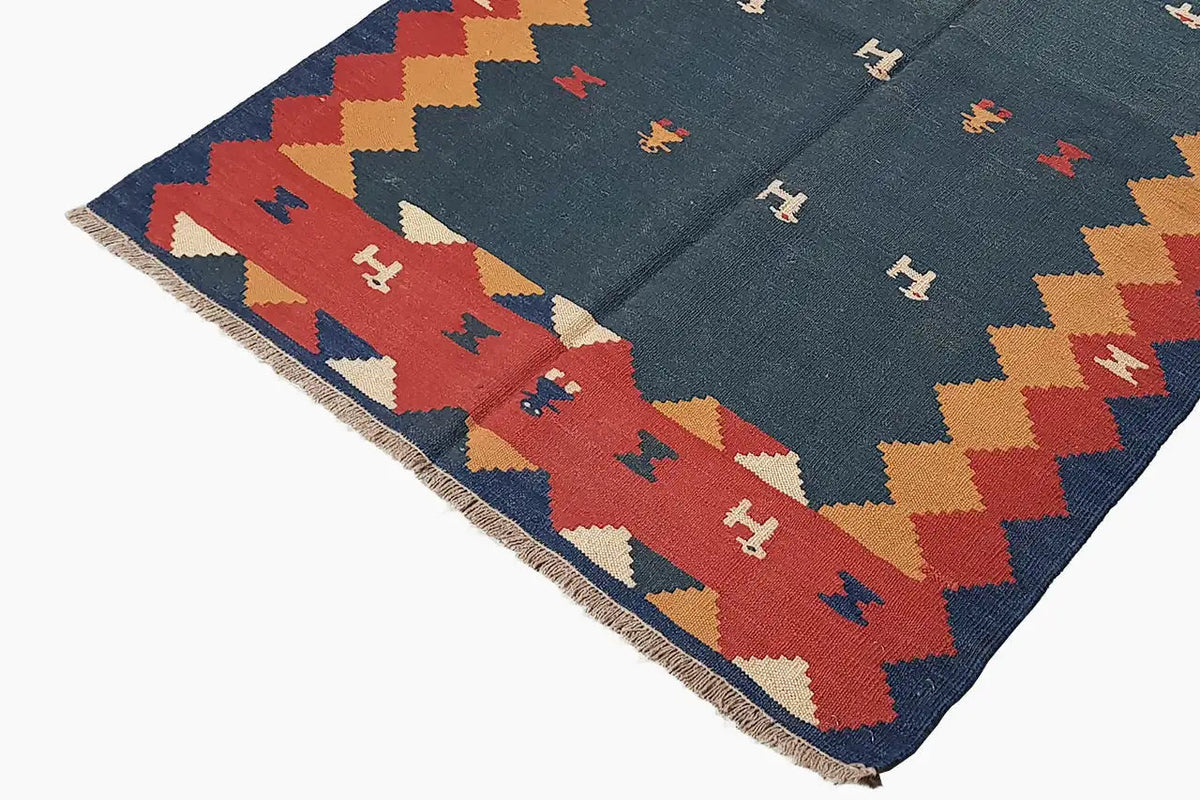 Kilim rug, photographed at an angle: blue patterned field with a zigzag border and animal/symbol motifs