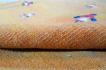 Close-up of carpet fabric: warm brown-orange, coarse weave, and occasional colorful threads.