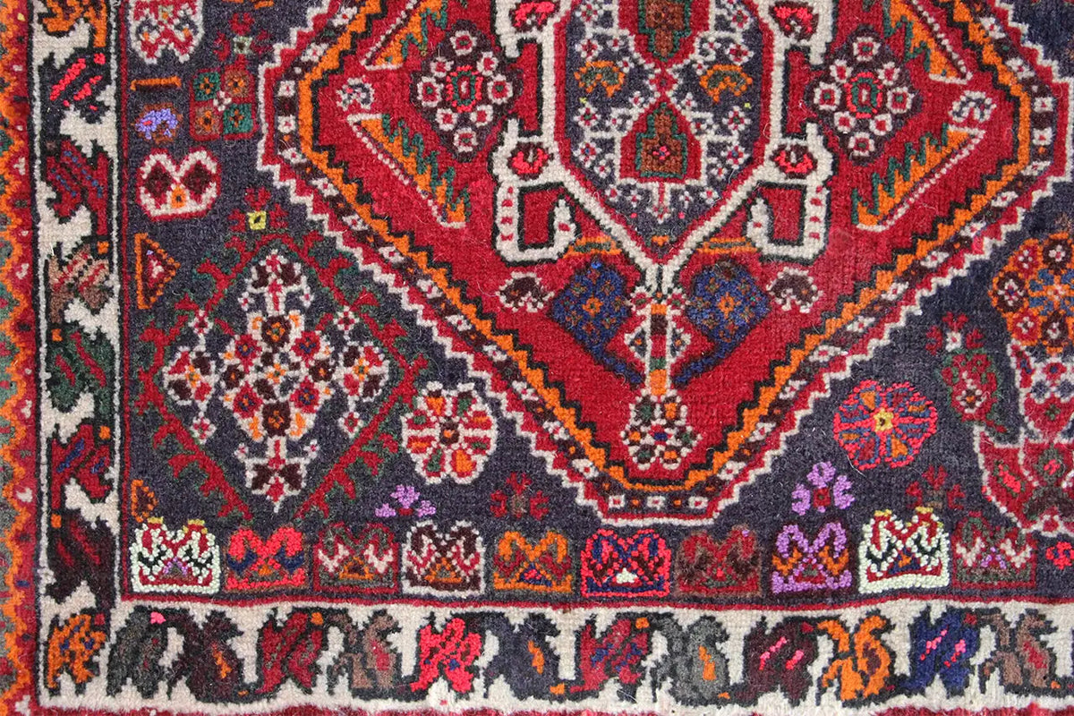 Close-up of a carpet: dense ornamental pattern in red, blue, and gray with a colorful border at the bottom.