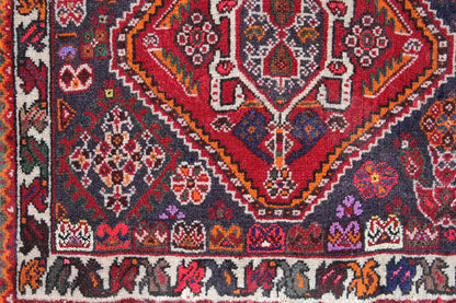 Close-up of a carpet: dense ornamental pattern in red, blue, and gray with a colorful border at the bottom.