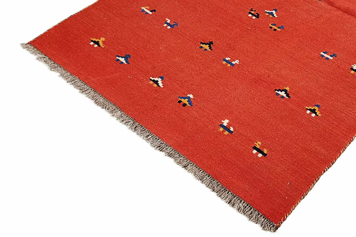 Red kilim rug at an angle, with small colorful patterns and a frayed edge