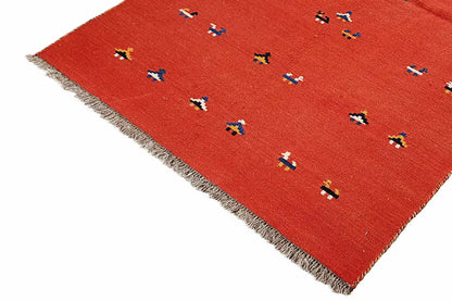 Red kilim rug at an angle, with small colorful patterns and a frayed edge
